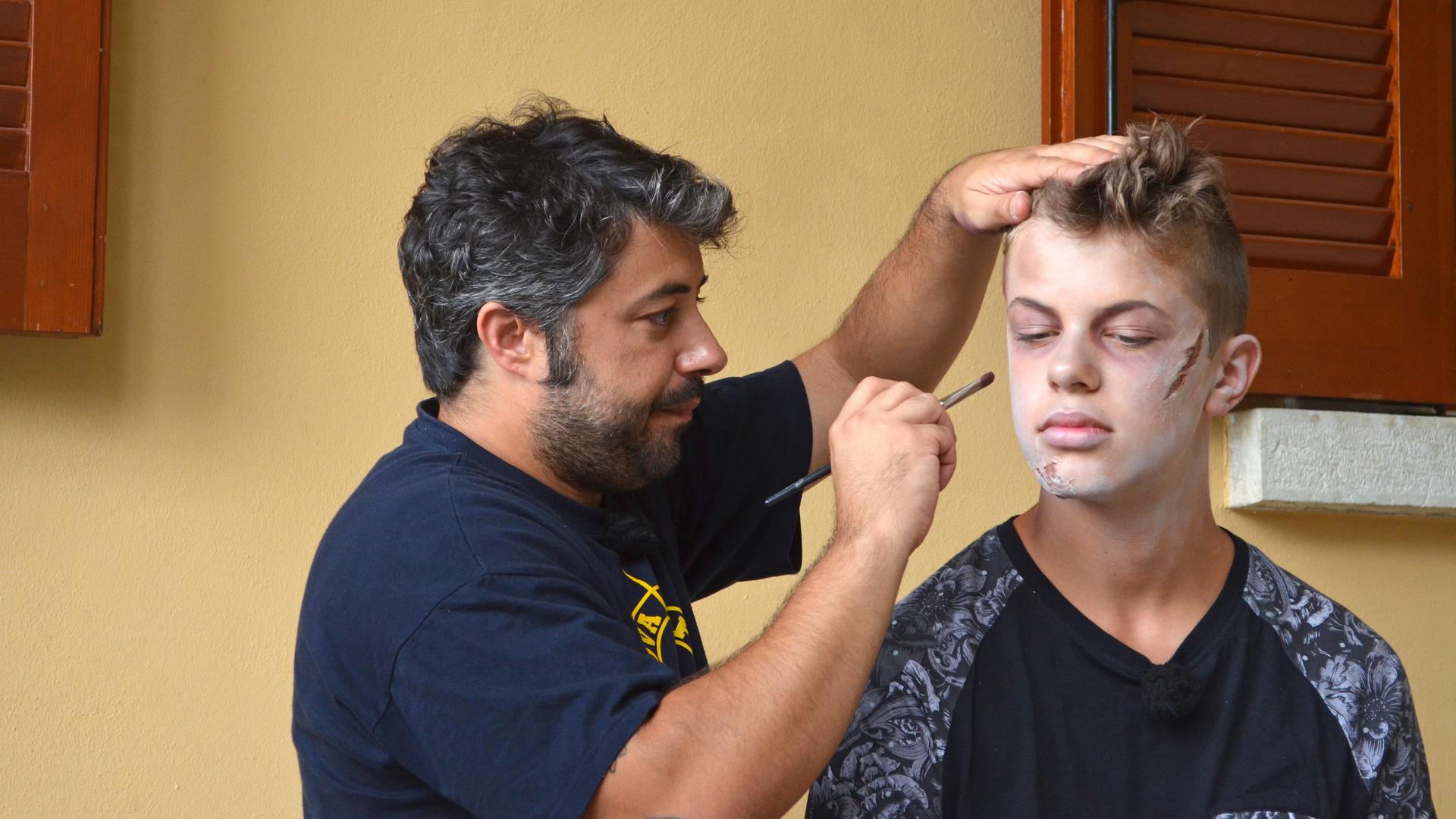 Einer der Jungs bekommt von einem Maskenbilder MakeUp aufgetragen, das ihn blass aussehen lässt und als hätte er Verletzungen in Gesicht.