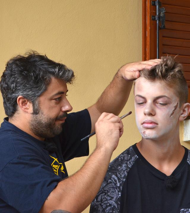 Einer der Jungs bekommt von einem Maskenbilder MakeUp aufgetragen, das ihn blass aussehen lässt und als hätte er Verletzungen in Gesicht.