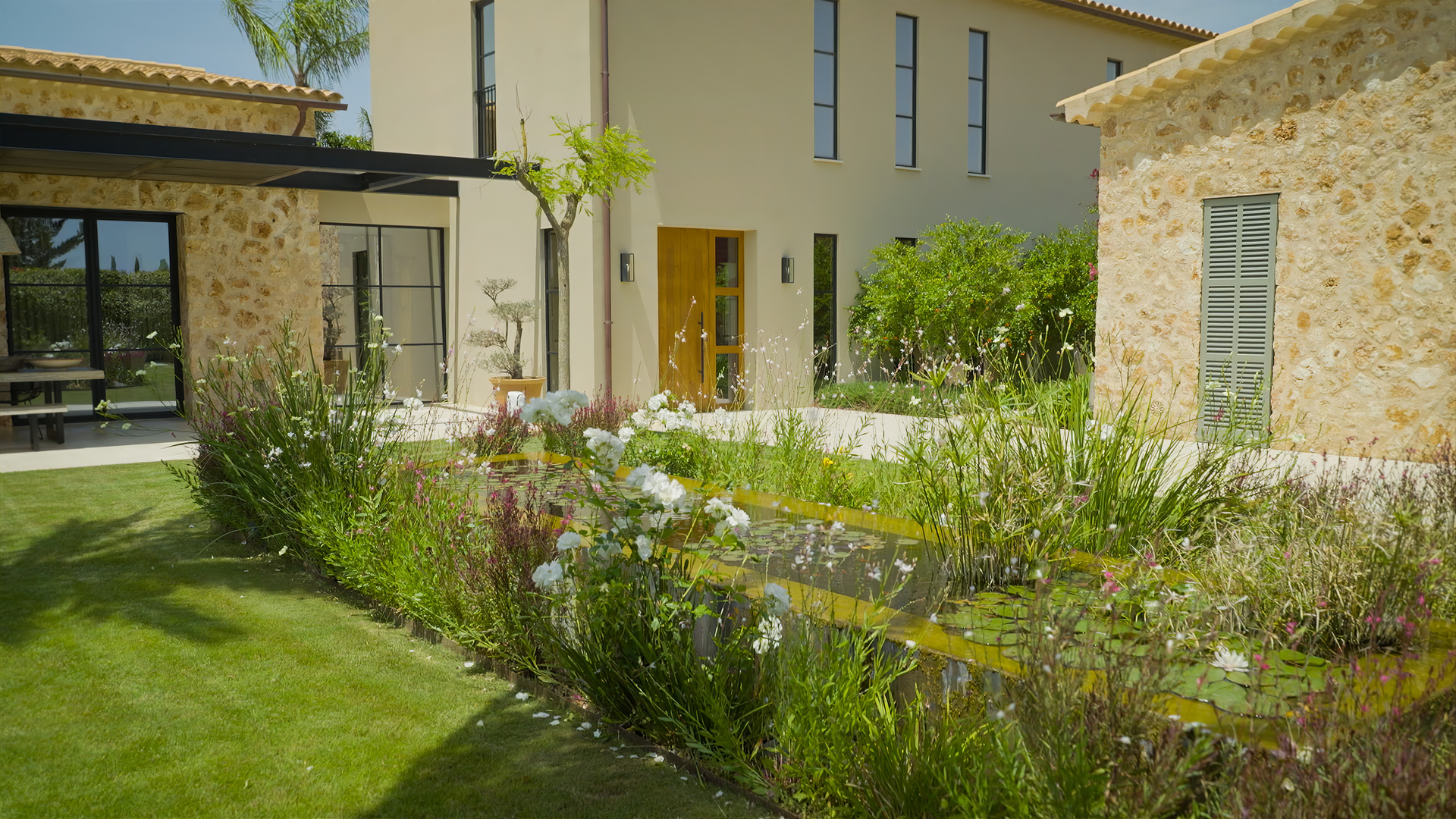 Folge 5: Garten mit Nachhaltigkeit: Eine Gartenfläche, von drei Gebäudefassaden umgeben, mit kurzgemähtem Rasen und einem von Blumen umgebenen Teich.