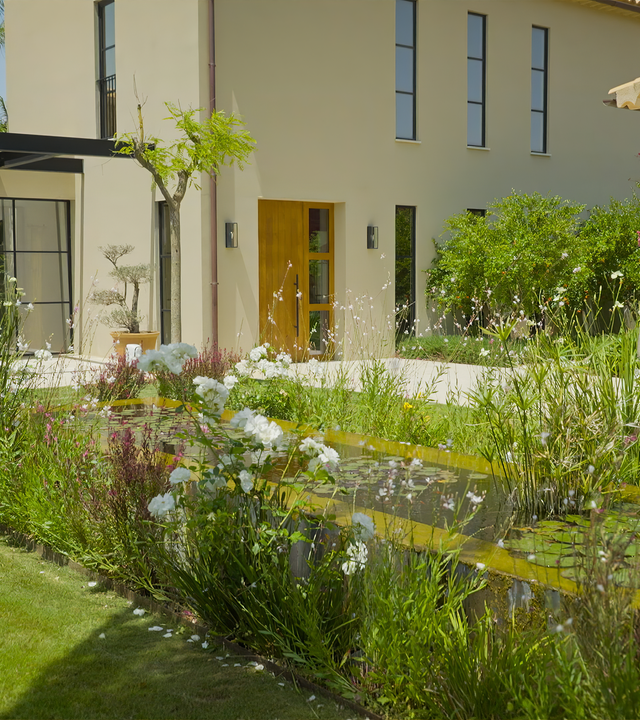 Folge 5: Garten mit Nachhaltigkeit: Eine Gartenfläche, von drei Gebäudefassaden umgeben, mit kurzgemähtem Rasen und einem von Blumen umgebenen Teich.