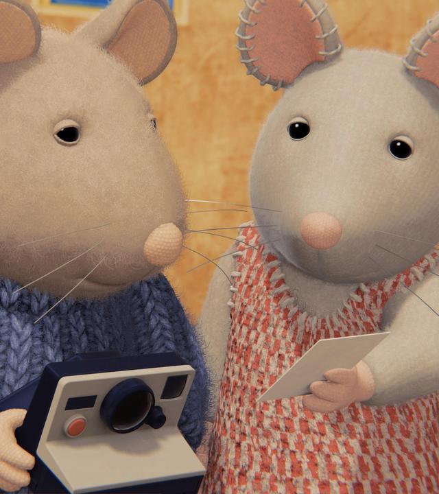 Sam und Julia stehen nebeneinander und schauen gemeinsam auf ein Foto, das Julia mit der linken Hand hochhält. Sam hält eine Polaroidkamera in beiden Händen.  Beide stehen in einem gepflasterten Hof vor einem Gebäude.