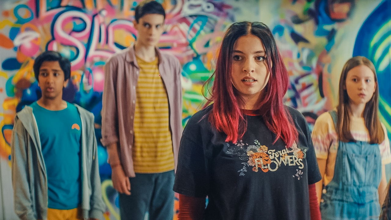 Von links nach rechts: Glen, Louis, Kaz und Meg (gespielt von Krish Misra, Oliver Cunliffe, Maiya Silveston und Katy Byrne) stehen vor einer bunten Graffitiwand und schauen überrascht. Neben ihnen steht Maeve, gespielt von Skye McClenaghan. Sie spricht zu ihnen.