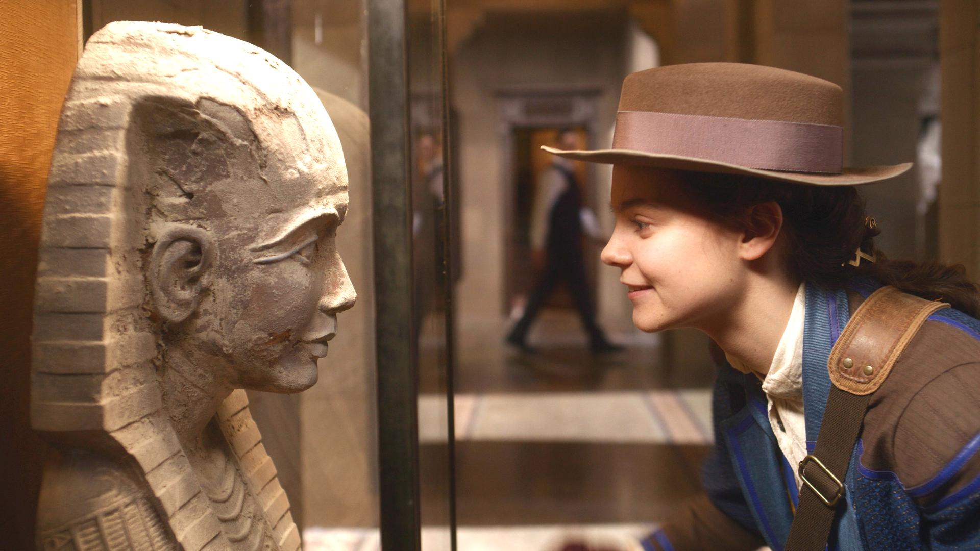 Im Museum: Theodosia schaut lächelnd und leicht zu ihr hin gebeugt einer steinernen Pharaonen-Statue in die Augen. Sie trägt einen braunen Hut.