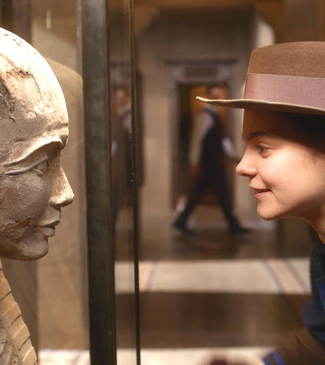 Im Museum: Theodosia schaut lächelnd und leicht zu ihr hin gebeugt einer steinernen Pharaonen-Statue in die Augen. Sie trägt einen braunen Hut.