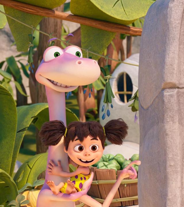 Der rosafarbene Dinosaurier Minus Drei hält seine Freundin, das Menschenmädchen Lucy, im Arm. Er zeigt auf eine steinzeitliche Maschine mit Holzknöpfen.