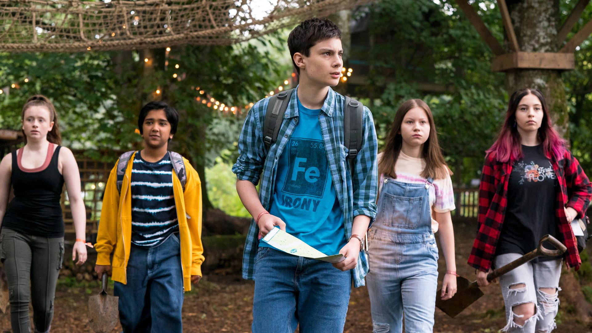 Im Wald vor einem hohen Klettergerüst aus Holz und einer Seilleiter mit Netzgeländer laufen fünf Jugendliche versetzt nebeneinander her - Bea (Aoife Hughes), Glen (Krish Misra), Louis (Oliver Cunliffe), Meg (Katy Byrne) und Kaz (Maiya Silveston). Louis in der Mitte hält einen Zettel in der Hand.