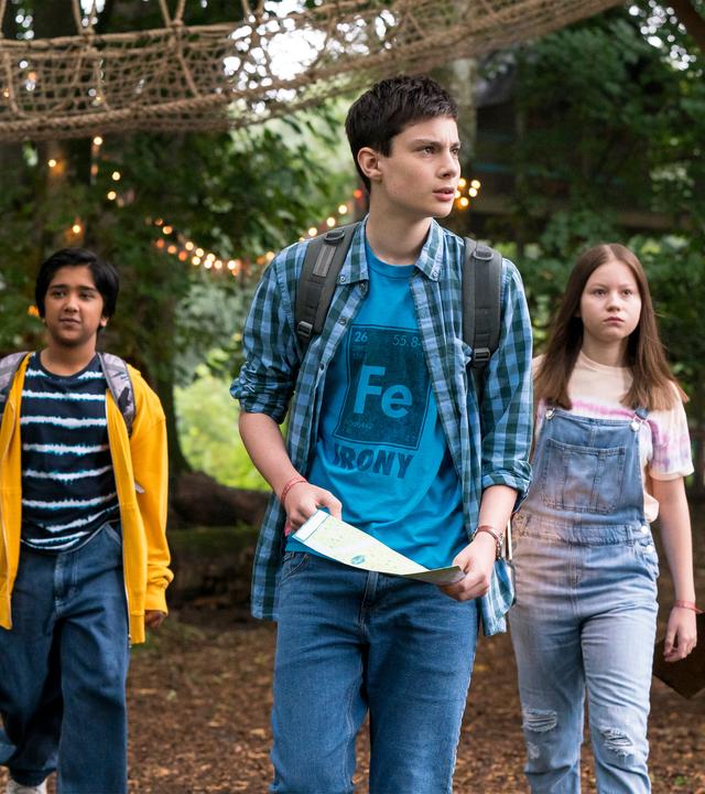 Im Wald vor einem hohen Klettergerüst aus Holz und einer Seilleiter mit Netzgeländer laufen fünf Jugendliche versetzt nebeneinander her - Bea (Aoife Hughes), Glen (Krish Misra), Louis (Oliver Cunliffe), Meg (Katy Byrne) und Kaz (Maiya Silveston). Louis in der Mitte hält einen Zettel in der Hand.