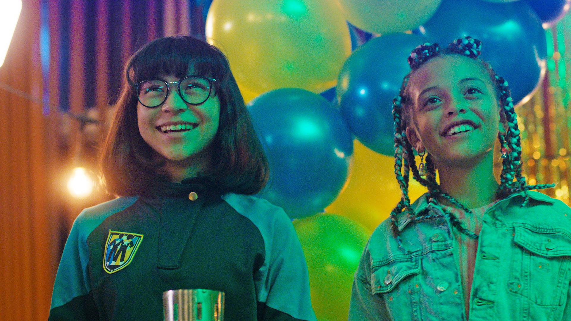 Die beiden Mädchen Alpha und Jenny stehen glücklich lächelnd und nach vorne blickend nebeneinander in der Schulaula. Im Hintergrund hängen blaue und gelbe Luftballons als Partydekoration.