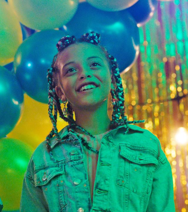 Die beiden Mädchen Alpha und Jenny stehen glücklich lächelnd und nach vorne blickend nebeneinander in der Schulaula. Im Hintergrund hängen blaue und gelbe Luftballons als Partydekoration.