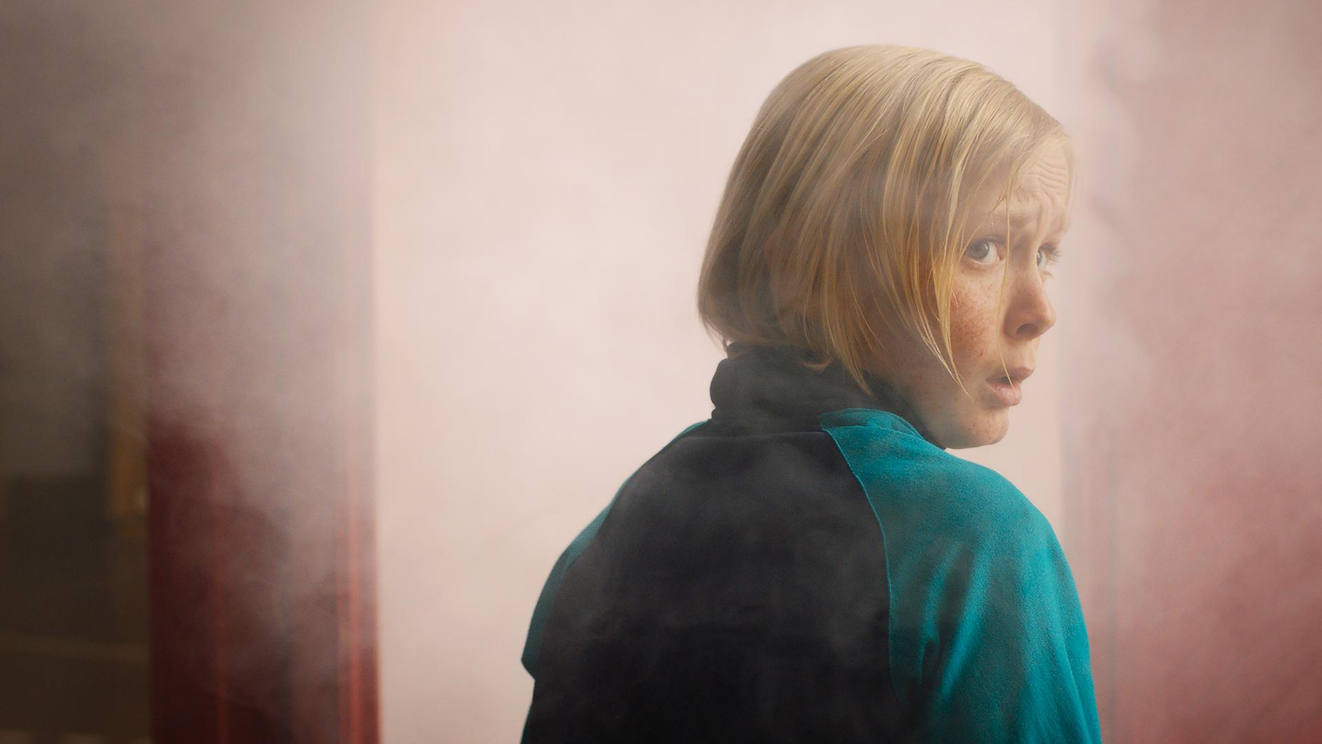 Raymond  schaut mit ängstlichem Blick über seine rechte Schulter nach hinten. Vor ihm ist eine geöffnete Tür zu sehen aus der Nebel steigt.