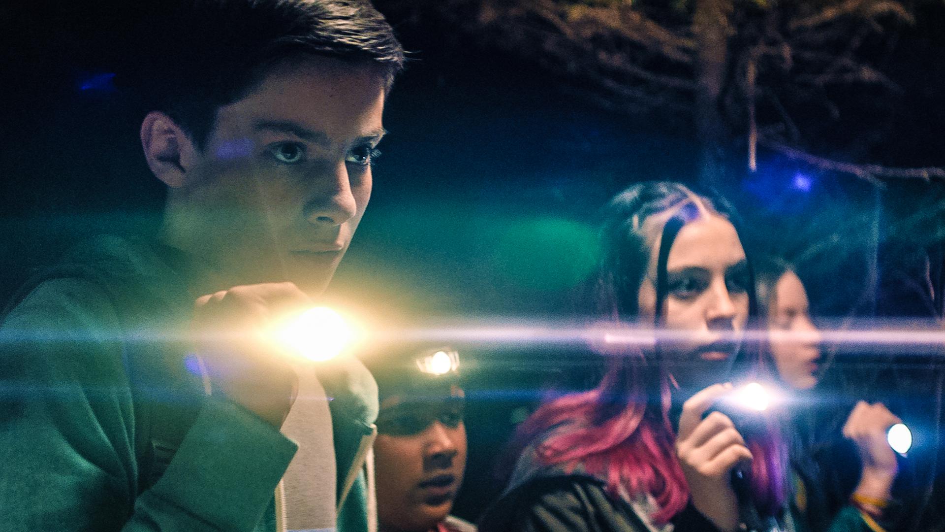 Im Wald bei Nacht stehen die vier Teenager der "Libellen"-Gruppe - Louis (Oliver Cunliffe), Glen (Krish Misra), Kaz (Maiya Silveston) und Meg (Katy Byrne) - mit Taschenlampen und einer Stirnlampe ausgerüstet in einer Reihe. Alle haben ernste Gesichtsausdrücke.