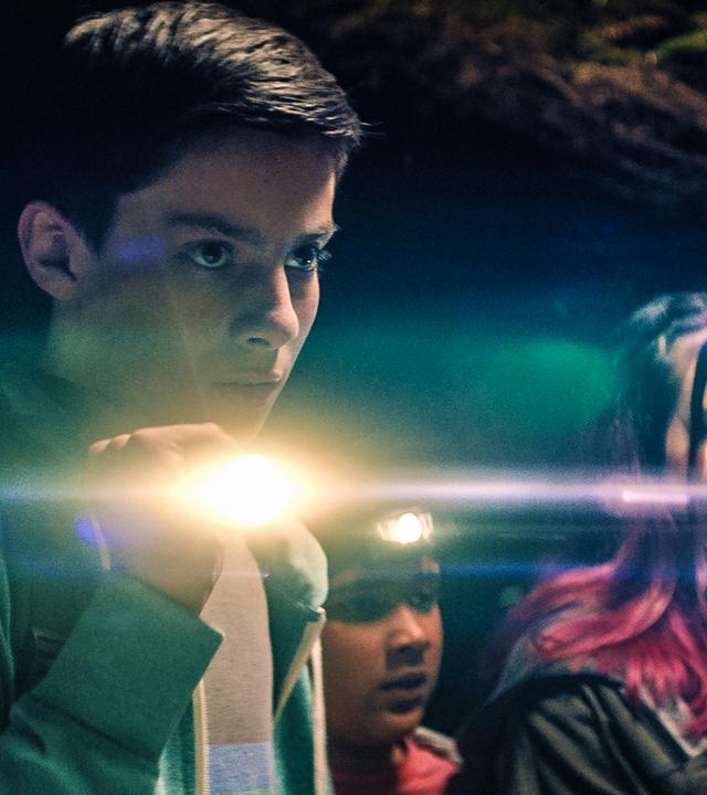 Im Wald bei Nacht stehen die vier Teenager der "Libellen"-Gruppe - Louis (Oliver Cunliffe), Glen (Krish Misra), Kaz (Maiya Silveston) und Meg (Katy Byrne) - mit Taschenlampen und einer Stirnlampe ausgerüstet in einer Reihe. Alle haben ernste Gesichtsausdrücke.
