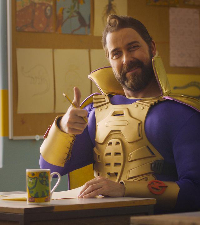 Thunderbolt sitzt an einem Schülerpult im Klassenzimmer. Er hält lächelnd seinen rechten Daumen nach oben und blickt nach vorne. Zwischen seinen Fingern klemmt ein Bleistift. Mit der linken Hand hält er ein Blatt Papier fest, das auf dem Tisch liegt. Vor ihm steht eine Tasse mit Katzenmotiv.