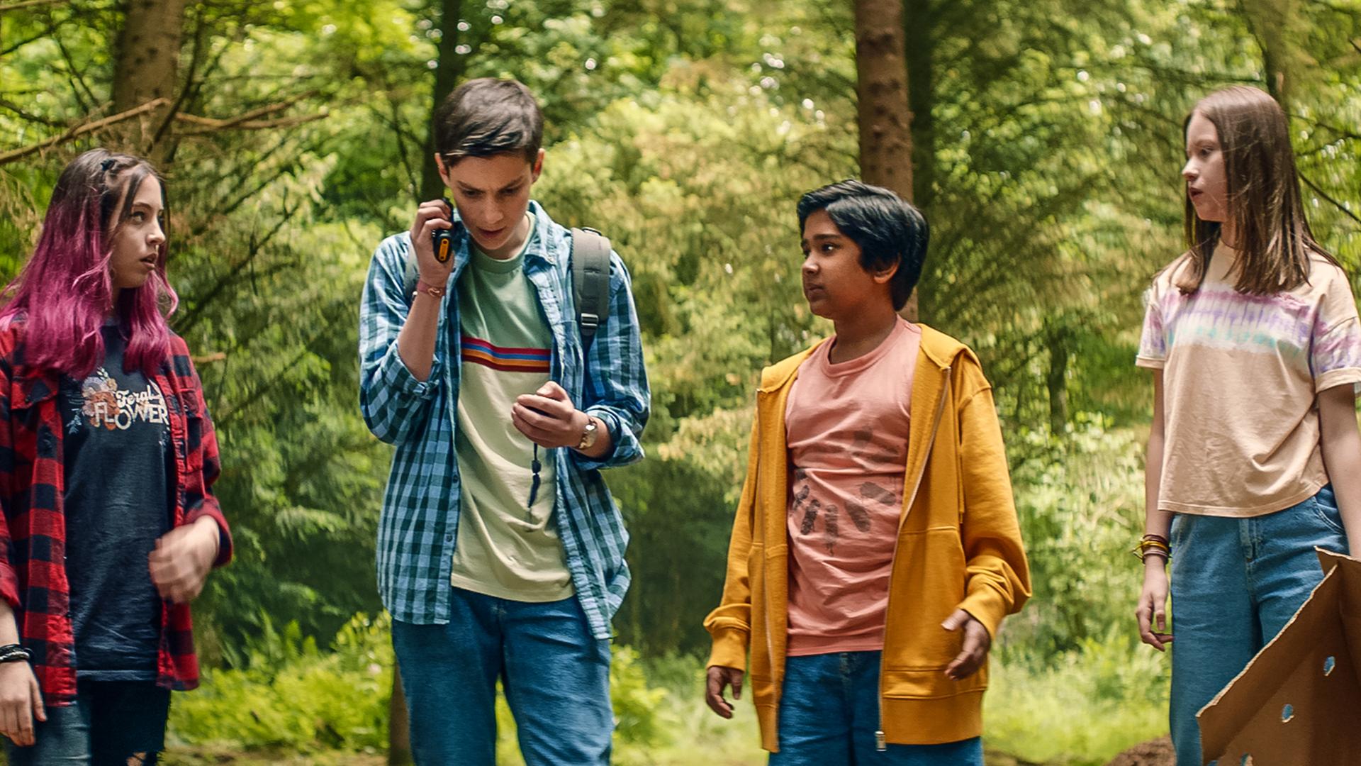 Im Wald auf einer Lichtung stehen Kaz, Louis, Glen und Meg nebeneinander. Kaz, Glen und Meg schauen gespannt zu Louis, der ein Walkie-Talkie am Ohr hat. Meg hält einen leeren offenen Karton in der Hand.