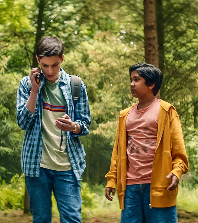 Im Wald auf einer Lichtung stehen Kaz, Louis, Glen und Meg nebeneinander. Kaz, Glen und Meg schauen gespannt zu Louis, der ein Walkie-Talkie am Ohr hat. Meg hält einen leeren offenen Karton in der Hand.