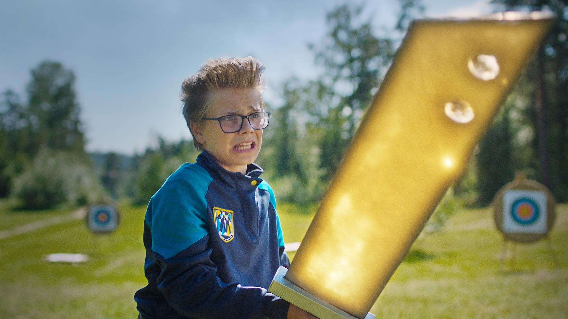 Uno steht mit ängstlichem Blick auf einer Wiese, die von Bäumen und Sträuchern umringt ist. Er hält angestrengt ein riesiges golden leuchtendes Schwert mit beiden Händen fest. Im Hintergrund stehen Zielscheiben.