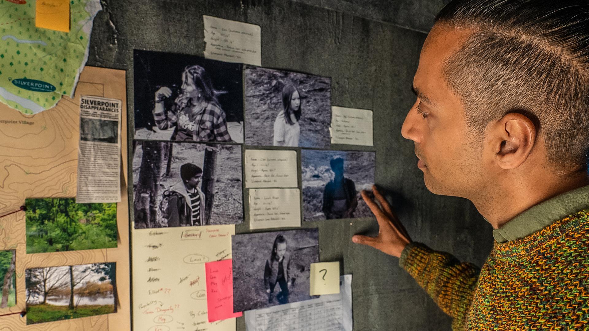 In einem Raum mit grauen Wänden steht der Mann namens Fischer vor verschiedenen Fotos, die angeleuchtet an der Wand hängen und schaut sie sich genau an. Um die Fotos herum hängen noch andere Zettel und eine Karte vom Camp Silverpoint.