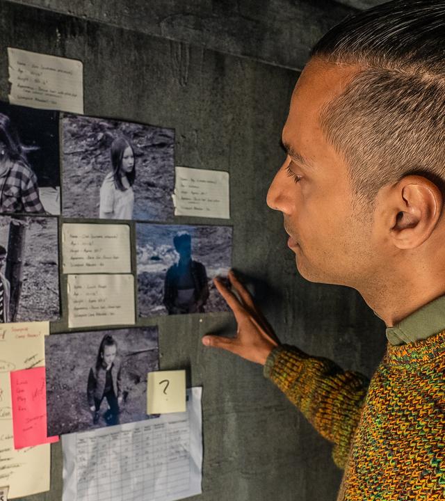In einem Raum mit grauen Wänden steht der Mann namens Fischer vor verschiedenen Fotos, die angeleuchtet an der Wand hängen und schaut sie sich genau an. Um die Fotos herum hängen noch andere Zettel und eine Karte vom Camp Silverpoint.