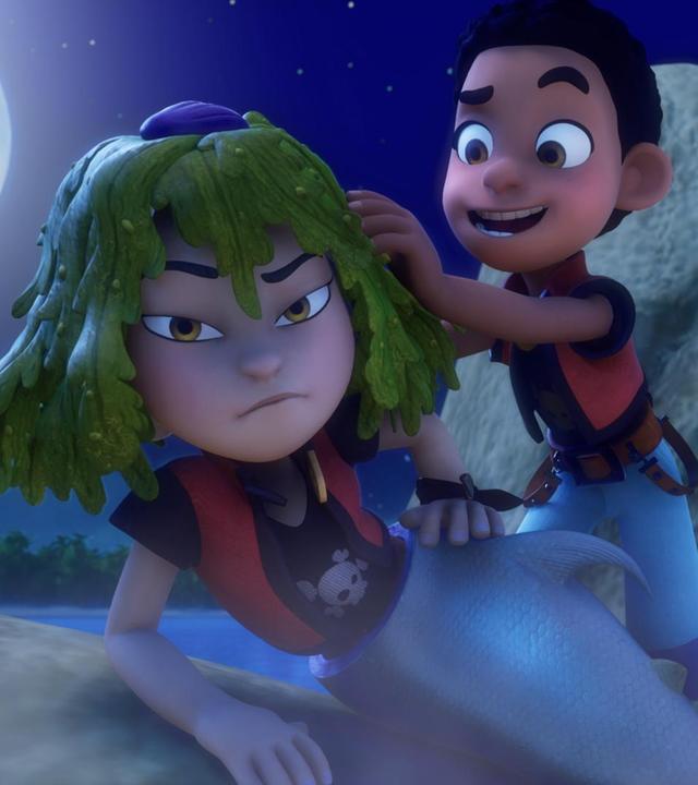 Finn steht nachts im Mondlicht auf einem Felsen hinter Zoe. Er fasst in ihr Algen-grünes Haar. Zoe hat eine Meerjungfrauenflosse an, stützt sich mit ihrem rechten Arm auf einem Felsen ab und hat ihren linken Arm in die Hüfte gestemmt. Ihr Gesichtsausdruck ist missmutig.