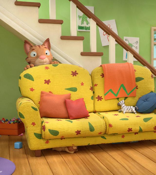 Im Wohnzimmer hinter dem bunten Sofa versteckt sich Frankie, die Katze. Vorsichtig schaut sie hinter der hohen Lehne des Sofas hervor. Im Hintergrund  hängen bunte, selbst gemalte Bilder am Treppenaufgang und durch ein Fenster blickt man hinaus in den Garten.