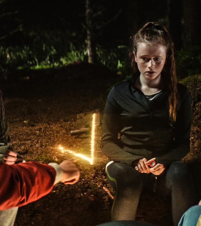 Im dunklen Wald auf der Lichtung sitzen Glen, Kaz, Bea und Louis mit ernsten Gesichtern um den Stein herum. Glen zeichnet sein Symbol, das als ein rechtwinkliger Lichtstrahl über dem Stein erscheint.