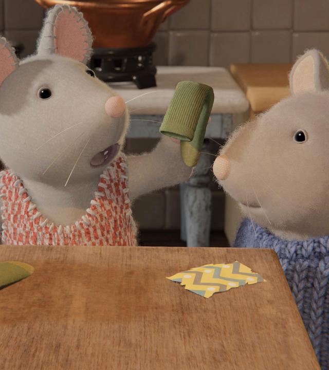 Julia und Sam stehen vor einem Holztisch in der Küche. Julia hält eine grüne Socke hoch, eine weitere liegt neben einem aufgerissenen Paket. Julia schaut enttäuscht, Sam etwas nachdenklich auf die Socke.