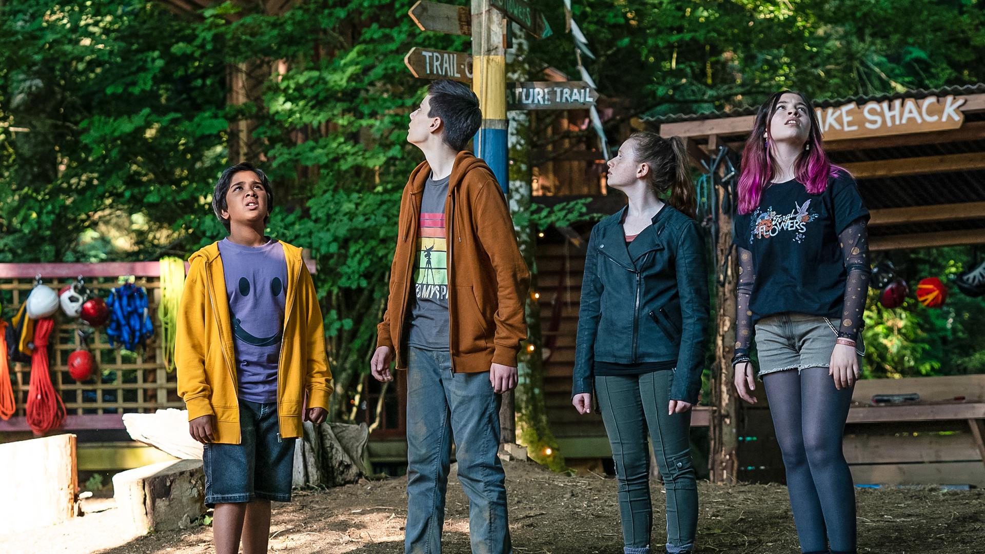 Tagsüber im menschenleeren Camp stehen Glen, Louis, Bea und Kaz und schauen suchend nach oben. Im Hintergrund stehen ein überdachter Holztisch, ein Holzmast und ein Holzgitter an dem Helme und Sportklamotten hängen.