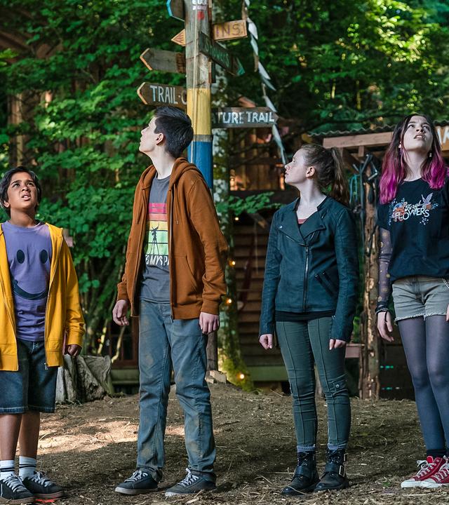Tagsüber im menschenleeren Camp stehen Glen, Louis, Bea und Kaz und schauen suchend nach oben. Im Hintergrund stehen ein überdachter Holztisch, ein Holzmast und ein Holzgitter an dem Helme und Sportklamotten hängen.