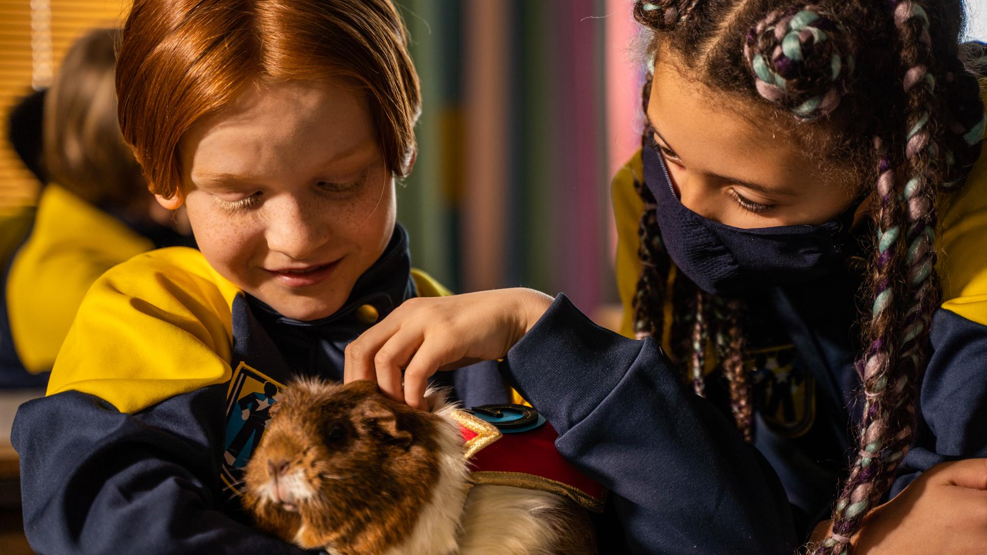 Magnus hält im Klassenzimmer glücklich lächelnd ein Meerschweinchen in seinen Armen und streichelt es mit seiner linken Hand. Rechts von ihm befindet sich Jenny, die sich mit einer Schutzmaske im Gesicht zum Meerschweinchen beugt.