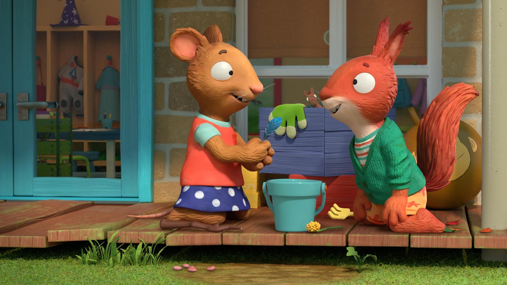 Tagsüber auf der Holzveranda des Hauses spielen Posy, die Maus und Jamila das Eichhörnchen  - sie brauen einen Zaubertrank. Beide knien vor einem Eimer und überlegen, welche Zutaten sie mixen können. Posy hält eine blaue Feder in der Hand und blickt zu Jamila, die zustimmend lächelt.