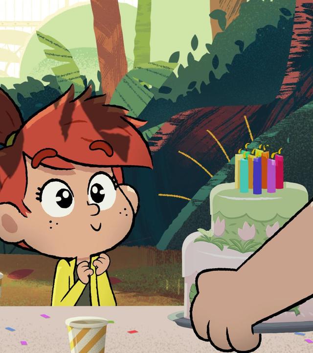 Ein Mädchen mit roten Haaren sitzt freudig aufgeregt im Dschungel an einem gedeckten Tisch und bekommt eine doppelstöckige Torte mit brennenden Kerzen gebracht.