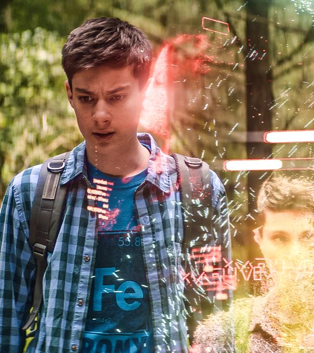 Im Wald bei Tageslicht steht Louis mit ernstem Gesichtsausdruck neben einem transparenten Band, in dem sein Hologramm zweimal deutlich zu sehen ist. Kaz und Megs Hologramme sind zu erahnen. Zwei weiße Fortschritts-Balken sind über Louis Kopf im transparenten Band.