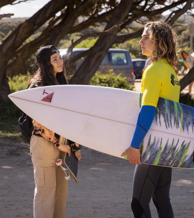 Das Mädchen Summer trifft zum ersten Mal den Sohn ihrer Gastfamilie, der mit dem Surfbrett unter dem Arm zum Strand läuft.