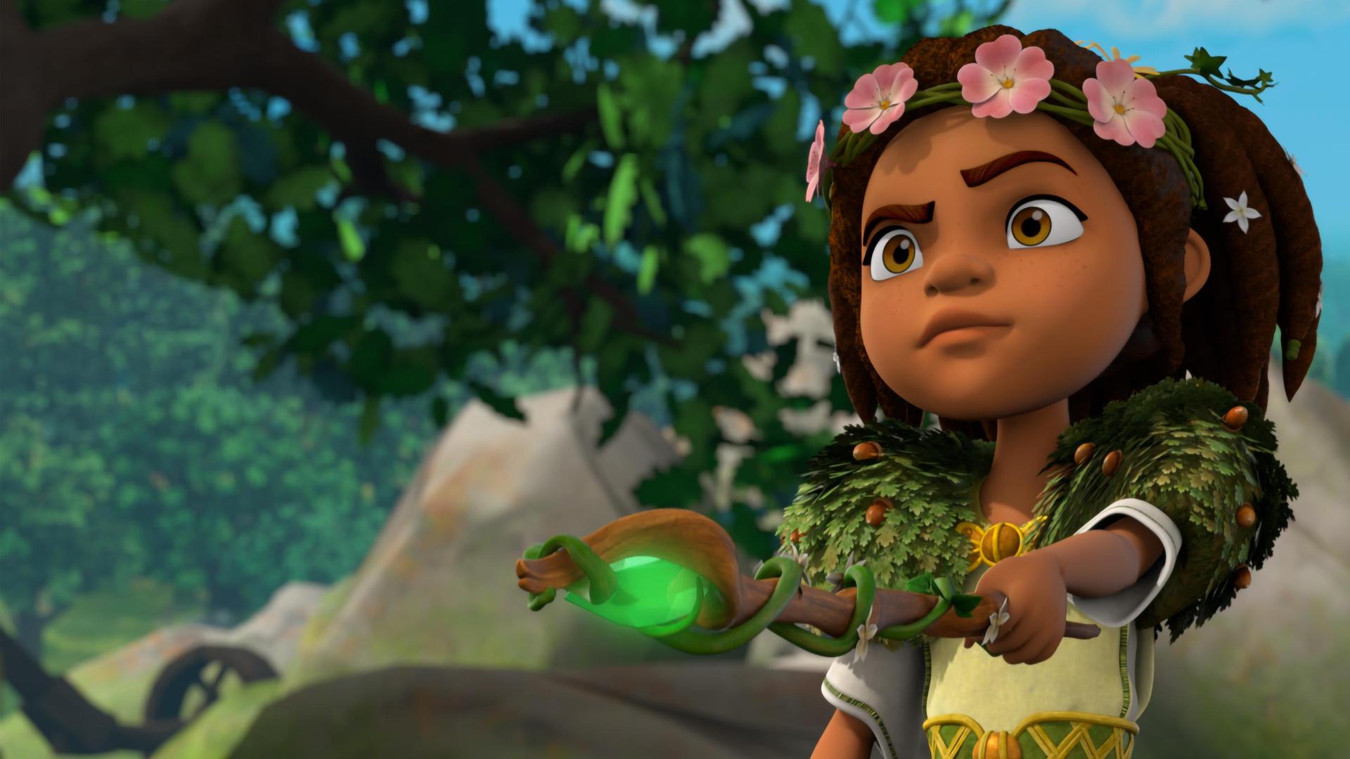 Im Wald vor großen Felsbrocken: Die Druidin Katell hält ihren Druidenstab senkrecht nach vorne auf ein Ziel gerichtet. Ein grüner, geschliffener Stein, der in die Spitze des Stabs eingebaut ist, leuchtet hell. Um ihren Kopf rankt ein Haarband, das mit rosa Blüten bestückt ist. Um ihre Schultern trägt sie eine Art Stola aus Eichenblättern und Eicheln und ihren Gürtel ziert eine Efeu-Schlinge.