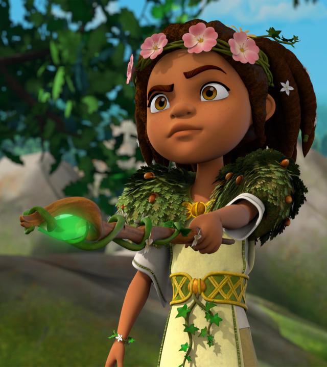 Im Wald vor großen Felsbrocken: Die Druidin Katell hält ihren Druidenstab senkrecht nach vorne auf ein Ziel gerichtet. Ein grüner, geschliffener Stein, der in die Spitze des Stabs eingebaut ist, leuchtet hell. Um ihren Kopf rankt ein Haarband, das mit rosa Blüten bestückt ist. Um ihre Schultern trägt sie eine Art Stola aus Eichenblättern und Eicheln und ihren Gürtel ziert eine Efeu-Schlinge.