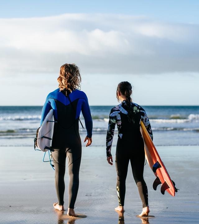 Ari und Summer gehen am Strand in Richtung Wasser. Sie sind von hinten zu sehen, tragen ihre Neoprenanzüge und jeder sein Surfbord unter dem Arm. Vor ihnen ist die Brandung.