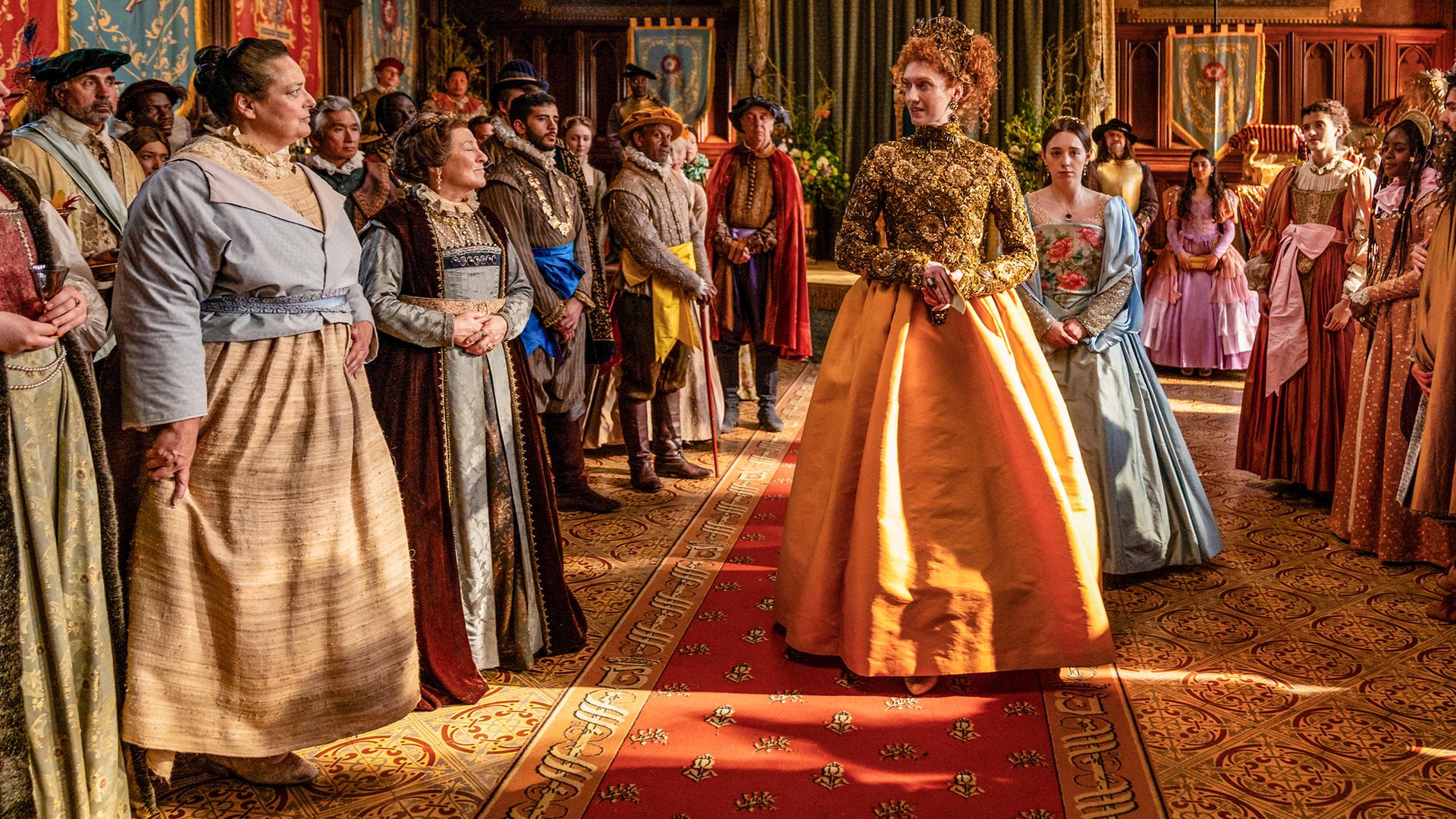 Die Queen läuft durch einen prunkvollen Saal auf einem Teppich durch die Menge ihrer Gäste. Viele Bedienstete, wie die Köchin und Mrs. Champernowne sind auch dabei.