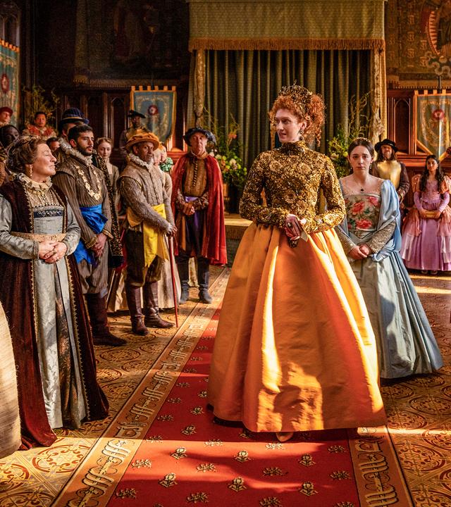 Die Queen läuft durch einen prunkvollen Saal auf einem Teppich durch die Menge ihrer Gäste. Viele Bedienstete, wie die Köchin und Mrs. Champernowne sind auch dabei.
