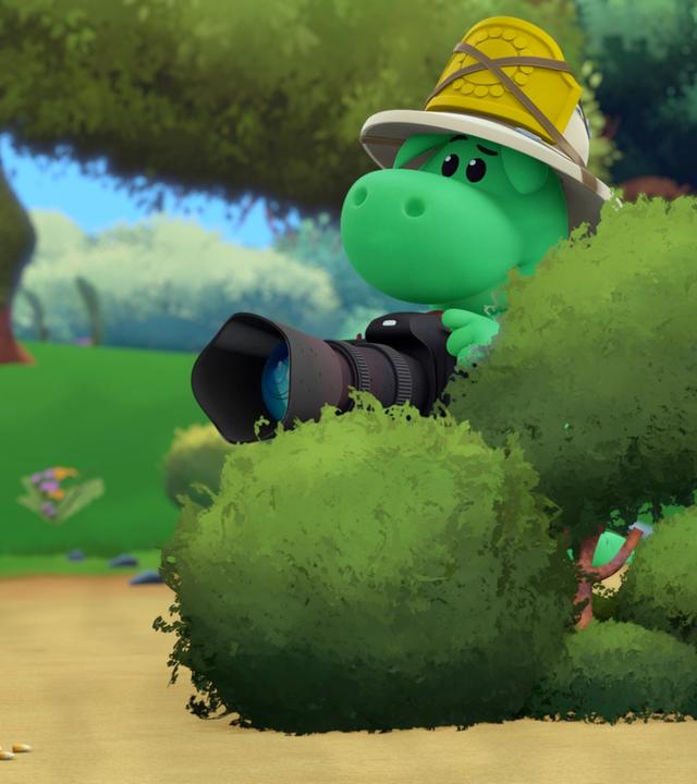 Grisu trägt einen Tropenhelm mit seinem Feuerwehrabzeichen darauf. Er versteckt sich mit Stella hinter einem Busch und hat, wie ein Tierfotograf, eine große Kamera in der Hand.