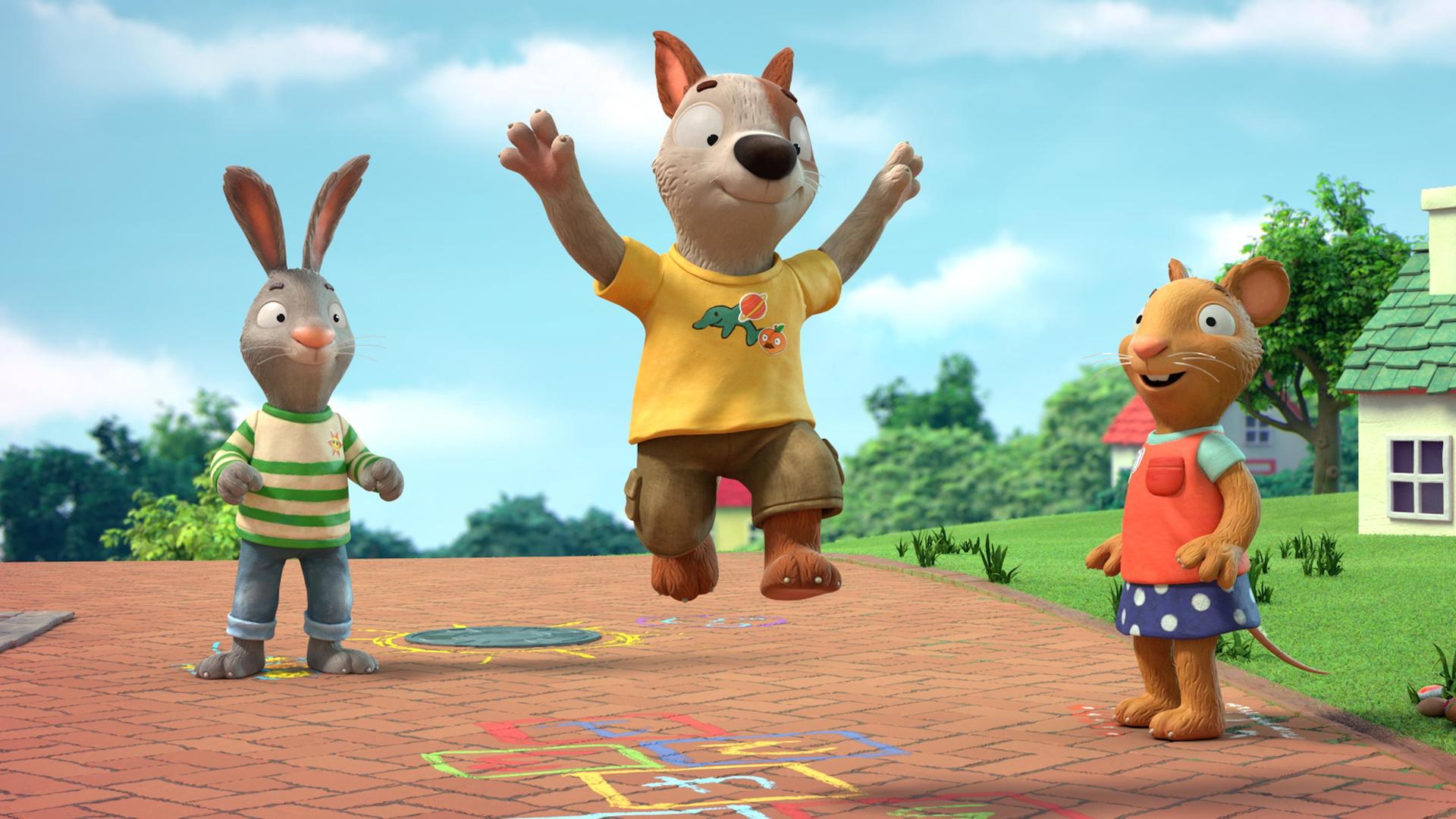 Auf einem gepflasterten Weg in einer Wohngegend zwischen Häusern spielen Pip, das Kaninchen, Posy, die Maus und Zac der Hund. Zac ist mitten im Spiel und gerade im Sprung. Mit bunter Kreide ist das Hüpfspiel Himmel und Hölle auf den Weg gemalt.