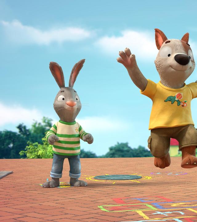 Auf einem gepflasterten Weg in einer Wohngegend zwischen Häusern spielen Pip, das Kaninchen, Posy, die Maus und Zac der Hund. Zac ist mitten im Spiel und gerade im Sprung. Mit bunter Kreide ist das Hüpfspiel Himmel und Hölle auf den Weg gemalt.