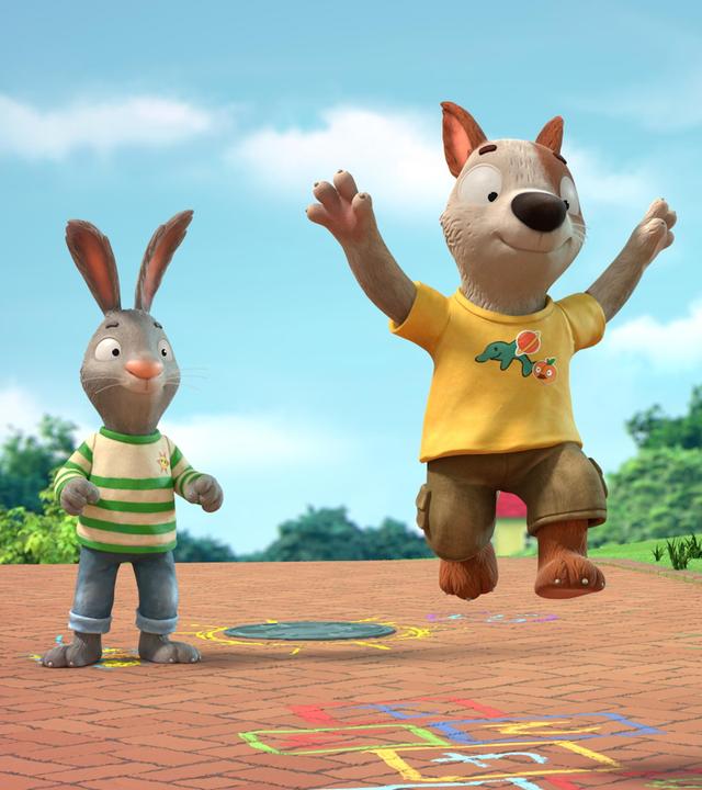 Auf einem gepflasterten Weg in einer Wohngegend zwischen Häusern spielen Pip, das Kaninchen, Posy, die Maus und Zac der Hund. Zac ist mitten im Spiel und gerade im Sprung. Mit bunter Kreide ist das Hüpfspiel Himmel und Hölle auf den Weg gemalt.