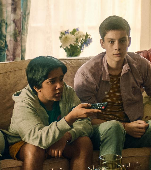 Kaz, Glen und Louis sitzen nebeneinander auf einem Sofa in einem Wohnzimmer. Kaz und Louis schauen erwartungsvoll nach vorne, während Glen mit einer Fernbedienung in seiner Hand ernst zur Seite schaut.
