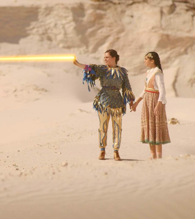 Theo und Safiya stehen Hand in Hand inmitten einer Sandlandschaft. Theo hat ihren rechten Arm ausgestreckt, von dem aus ein gelb leuchtender Strahl gesendet wird.