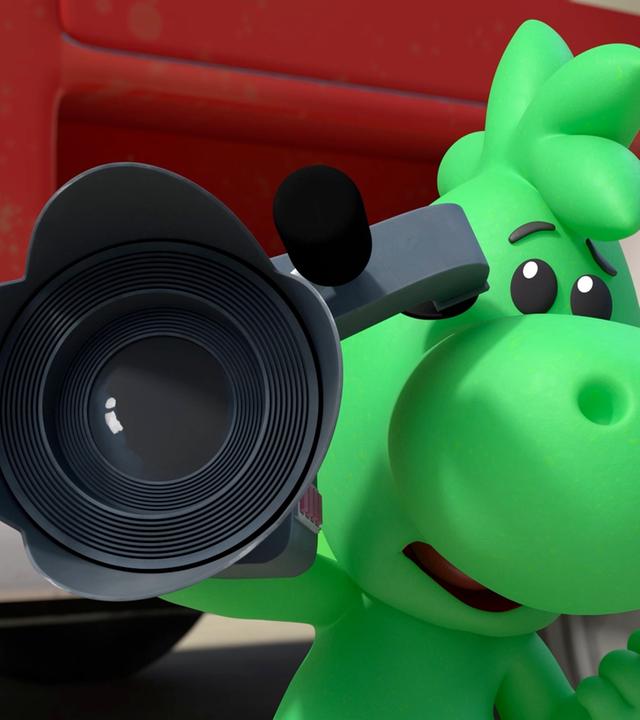 Grisu, der kleine grüne Drache, hält eine große Filmkamera in der Hand. Er steht vor einem Feuerwehrauto.