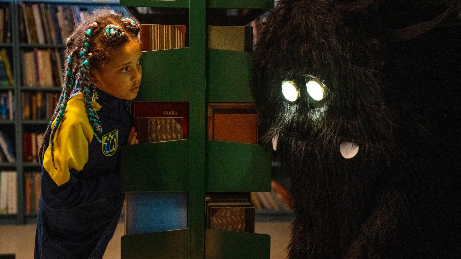 In der Bibliothek der Schule schaut Jenny an ein Bücherregal gelehnt und mit ungläubigem Blick auf ein zotteliges Wesen mit leuchtenden Augen, das vor ihr steht.