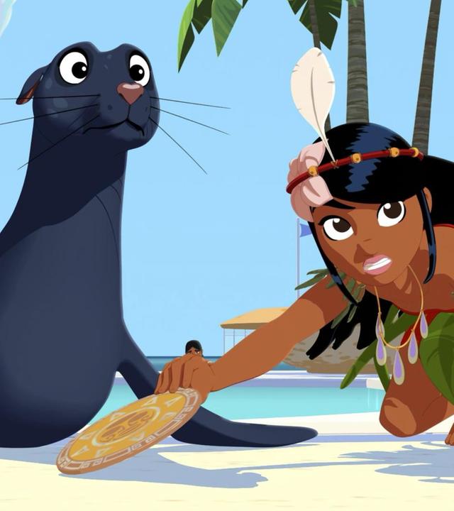 Am Strand liegt Seehündin Daisy neben Timeti, die auf dem Sand kniet und sich nach einer goldenen Scheibe streckt, die sie mit der rechten Hand berührt. Beide schauen nach vorne.