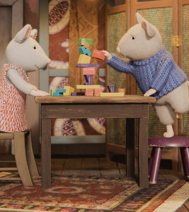 Julia kniet auf einem Stuhl an einem Tisch, Sam steht auf einem Hocker an diesem Tisch. Sam stellt gerade ein rotes Holzklötzchen auf einen Turm aus bunten Holzklötzchen. Julia schaut ihm aufmerksam dabei zu. Die beiden befinden sich in einem gemütlichen Zimmer des Mäusehauses. Rechts steht ein Bett hinter einem Paravent, links befindet sich eine Küchenzeile. Im Hintergrund sind eine Tür und ein Schrank zu sehen.