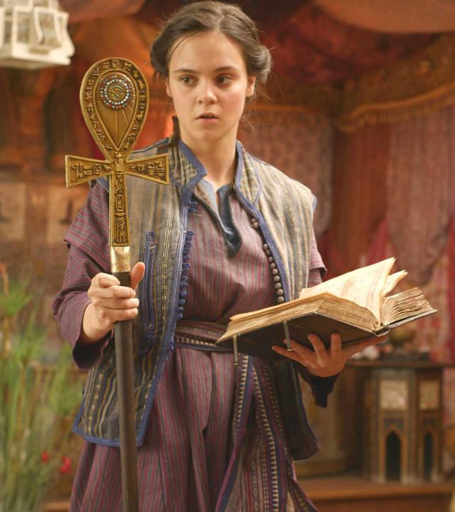 Theodosia hält in ihrer linken Hand ein aufgeschlagenes Buch und in der rechten einen Stab mit einem ägyptischen Symbol. Sie schaut den Stab mit erstauntem Blick und offenem Mund an. Neben ihr, auf dem Boden sitzend, befindet sich Safiya, die mit gespanntem Blick zu ihrer Freundin schaut.