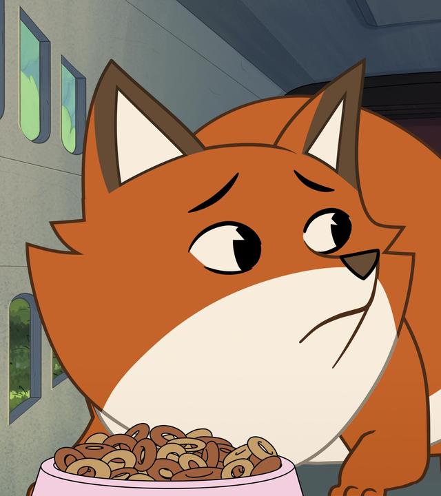 Der orangefarbene Fuchs Iwo ist in einer Transportkiste für Katzen eingesperrt. Die Kiste hat an der Seite kleine Luftschlitze. Iwo duckt sich mit ängstlichem Blick auf den Boden der kleinen Box. Vor ihm steht eine rosafarbene Schüssel mit Trockenfutter in Form von kleinen braunen Kringeln.